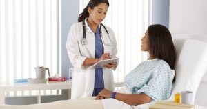 A woman in a hospital bed talks to a doctor holding a clipboard