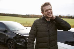a man holds his neck with two crashed cars in the background. 