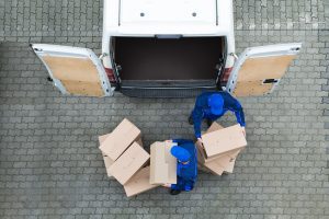 2 employees in blue overalls and caps unloading boxes out of the back of a white van. 