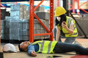 A warehouse worker checking on their colleague who is lying on the floor.