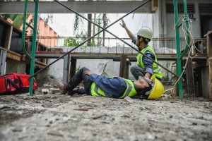 A construction worker laying on his back in pain, with another construction worker knelt beside him calling for assistance 