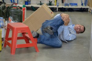 A man on the ground of a warehouse holding his knee lying next to a red stool.