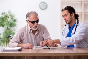 A man with severe vision loss wears sunglasses while learning braille with a doctor.