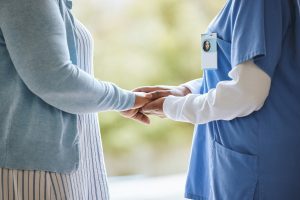 A carer holds the hands of an elderly client, both are standing upright against a blurred outdoor background. 